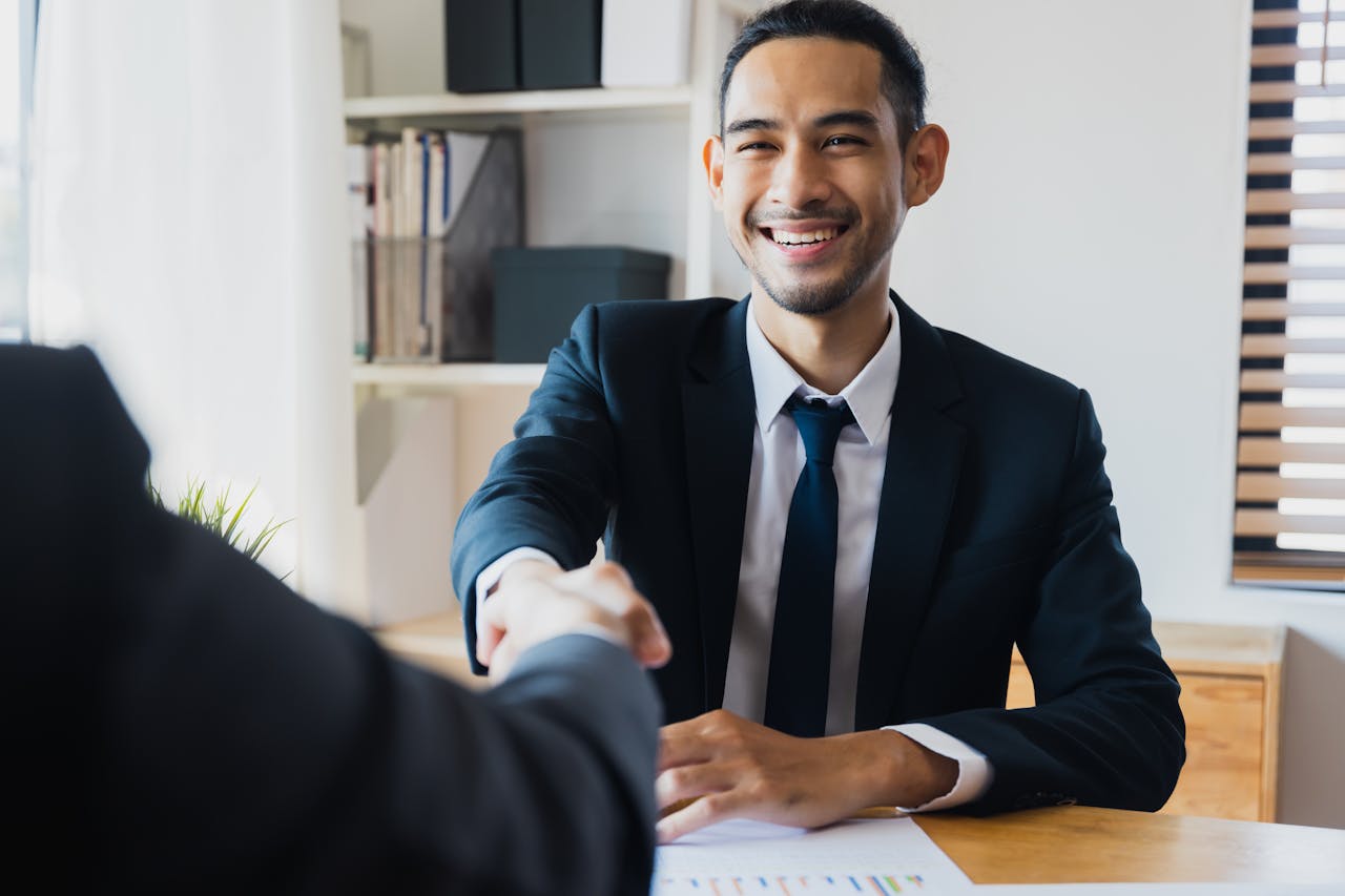 creative-02 Confident businessman in suit shaking hands at office desk, symbolizing successful partnership.