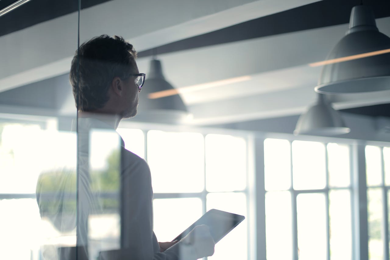 brand-02 A businessman holding a tablet in an office, looking thoughtfully through a glass wall.